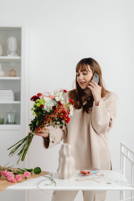 A woman with long brown hair and bangs, dressed in a beige sweater, is smiling and talking on a mobile phone while holding a large, vibrant bouquet of mixed fresh flowers including white roses, red ch
