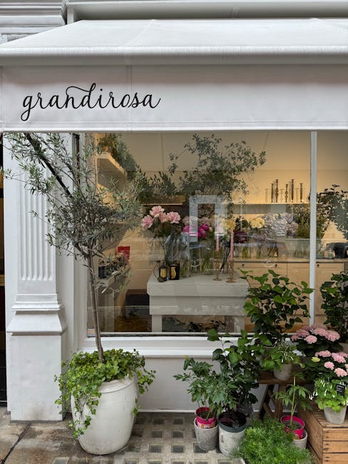 The image shows the exterior of a flower shop named 'grandirosa' with a white canopy and large glass window displaying an indoor floral arrangement. Outside, there are several potted plants and flower