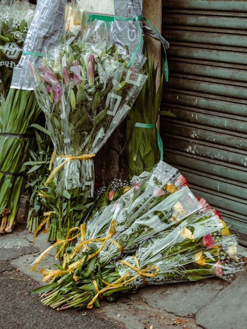 A collection of fresh, vibrant flower bouquets resting against a weathered wooden shutter on a pavement. The bouquets feature a variety of flowers, including pink lilies, yellow roses, and greenery, a