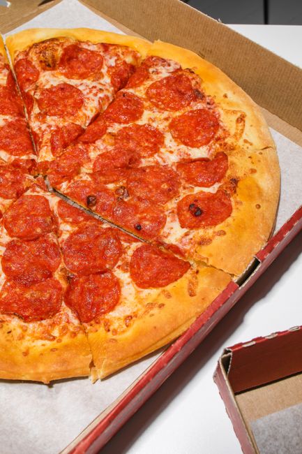 A close-up of a freshly baked pepperoni pizza inside an open cardboard delivery box. The pizza has a golden crust with a slightly crispy edge, topped with melted cheese and evenly spaced slices of spi