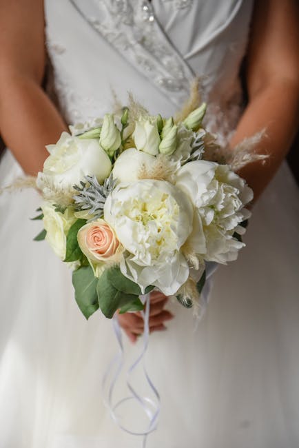 A bridal bouquet featuring predominantly white flowers, including large peonies, roses, and lisianthus, accented with soft gray foliage and delicate feather-like textures. The bouquet is arranged in a
