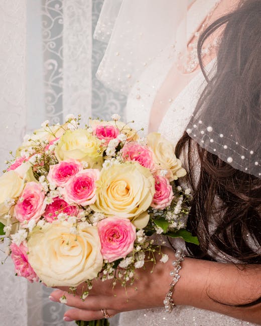 A woman in a bridal gown holds a floral bouquet arrangement consisting of large, creamy white roses and smaller pink roses, accented with delicate white baby's breath. The bouquet is tightly clustered