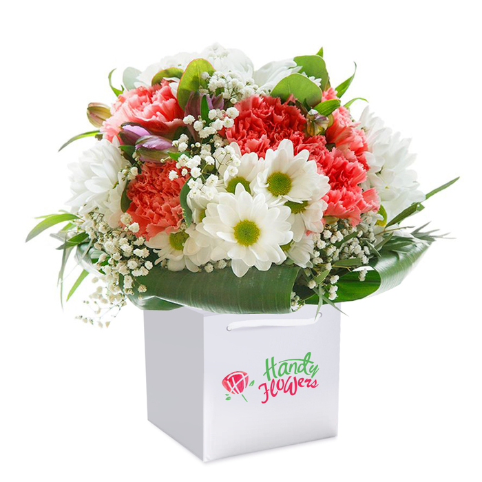 Upright in a pristine white square gift bag, this expertly crafted bouquet shines with the colours and textures that reflect Brondesbury's vibrant neighbourhood life. Soft coral, peach, and bold red carnations-each bloom flaunting lush, ruffled petals-create a richly textured canopy, punctuated by radiant white daisies with fresh lime-green centers. Gentle sprigs of baby's breath are scattered throughout, lending lightness and a hint of innocence, like blossoms glimpsed on a leisurely stroll through Mapesbury Dell. Along the left border, a few closed buds tinged with purple offer a subtle, mysterious charm, deepening the bouquet's visual appeal. An abundance of deep green, glossy leaves encircle the blooms, their broad forms elegantly framing the composition and underscoring the freshness synonymous with flower stands along Brondesbury Park. The bag's white ribbon handles suggest convenience without sacrificing style, ideal for a hand-delivered gesture to homes or celebrations throughout the area. Every element is thoughtfully placed, emphasising quality and the artistry of a florist who knows how to convey emotion through flowers-ready to uplift birthdays, anniversaries, or everyday moments across Brondesbury's welcoming streets.